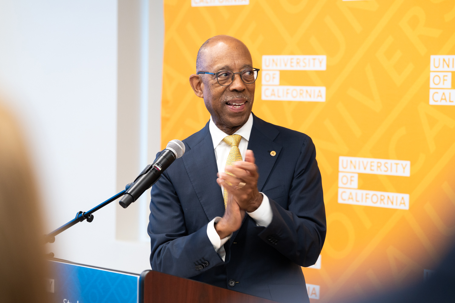 President Drake at a lectern, hands clasped and smiling