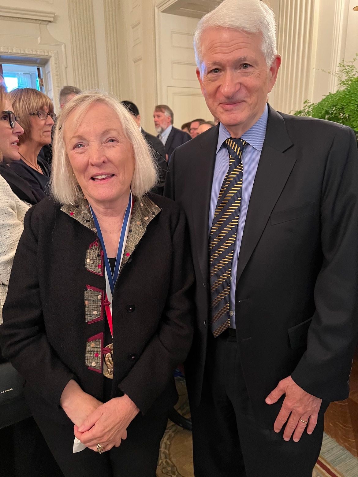 Shelley Taylor, white with a white/blonde bob haircut, wears a medal and smiles for the camera with UCLA Chancellor Gene Block, white with white hair, wearing a suit.