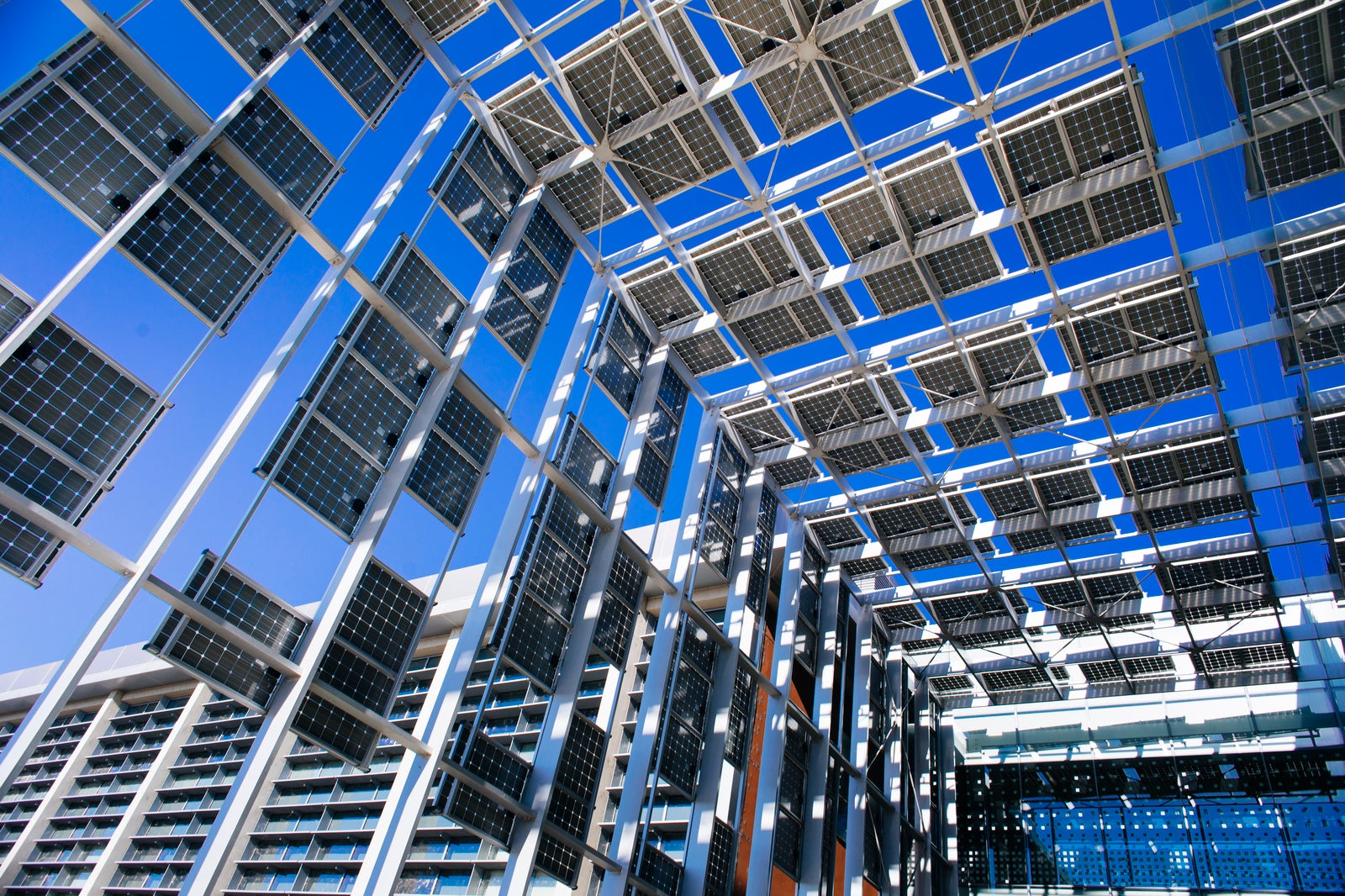 Looking up at a tall breezeway leading into a glass-walled building - the breezeway is constructed of solar panels on a scaffold. The sky is very blue.
