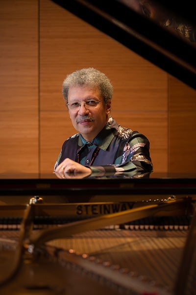 A Black man with gray hair, a moustache and glasses stands framed by a piano and its lid