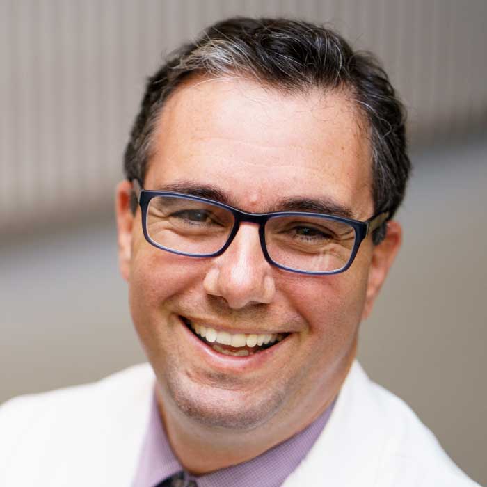 Man with short dark hair and glasses in a white coat, smiling