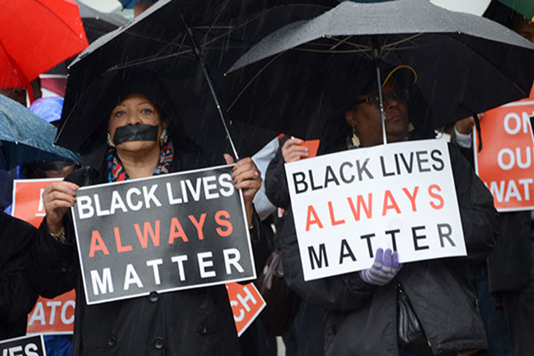 Black Lives Matter protesters with tape over their mouths stand with signs under umbrellas