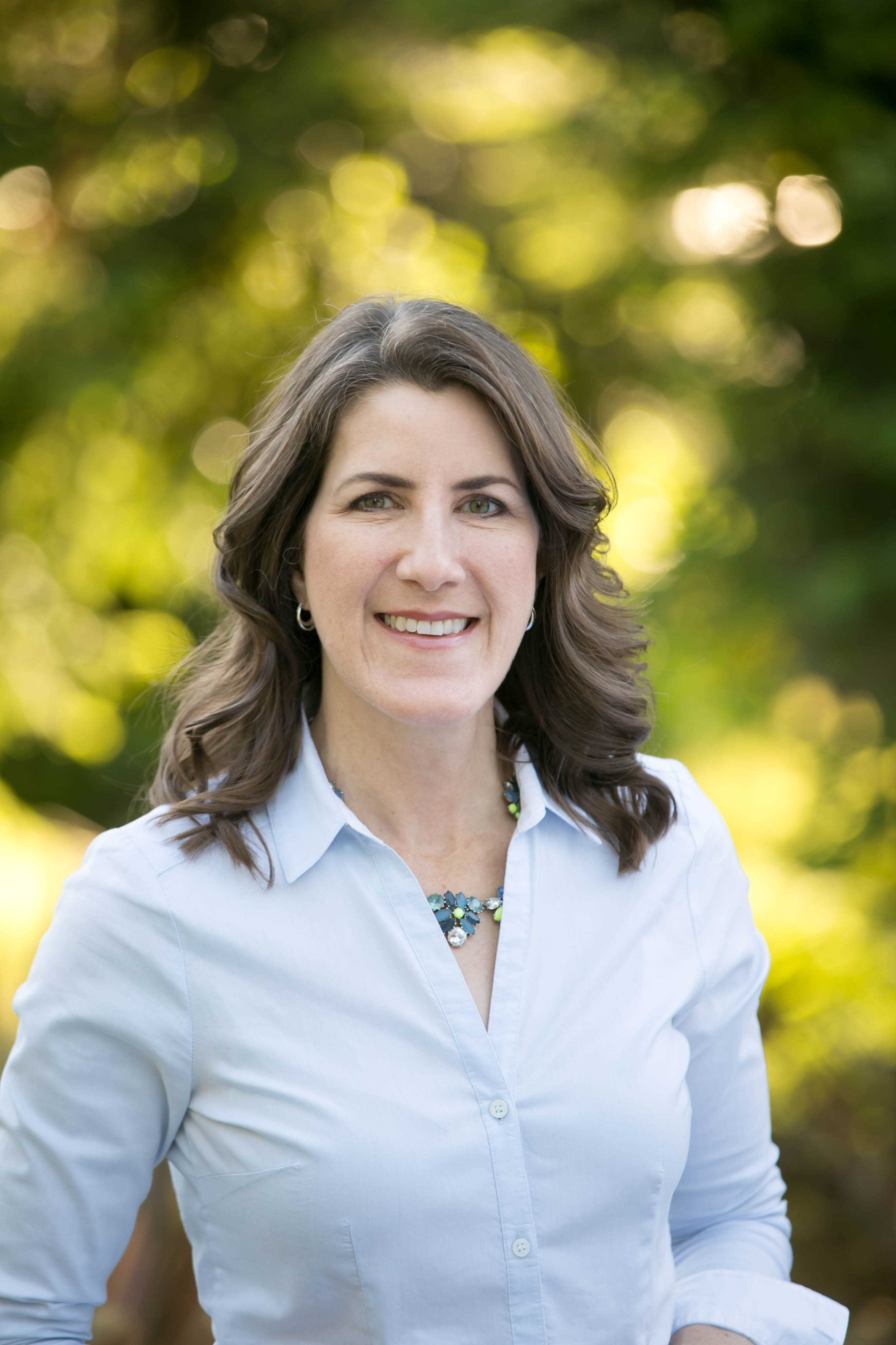 woman with shoulder-length wavy hair smiles in front of an out-of-focus outdoor background