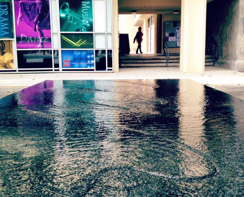 A flat fountain covered with water with curving linear patterns, with a breezeway and a person walking in the background