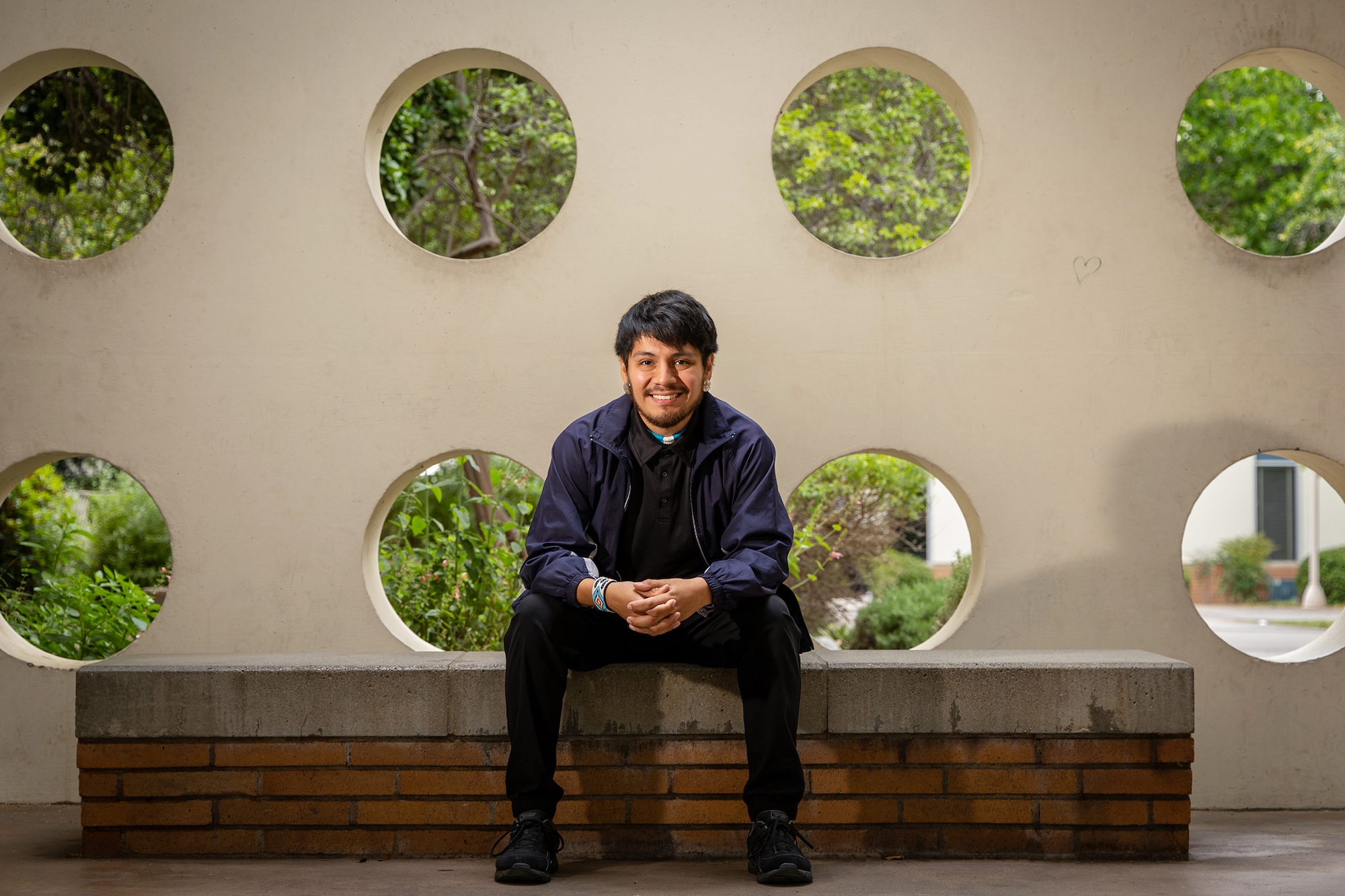 Young man with beard smiles in front of a wall with several circular cutouts, showing trees and greenery behind
