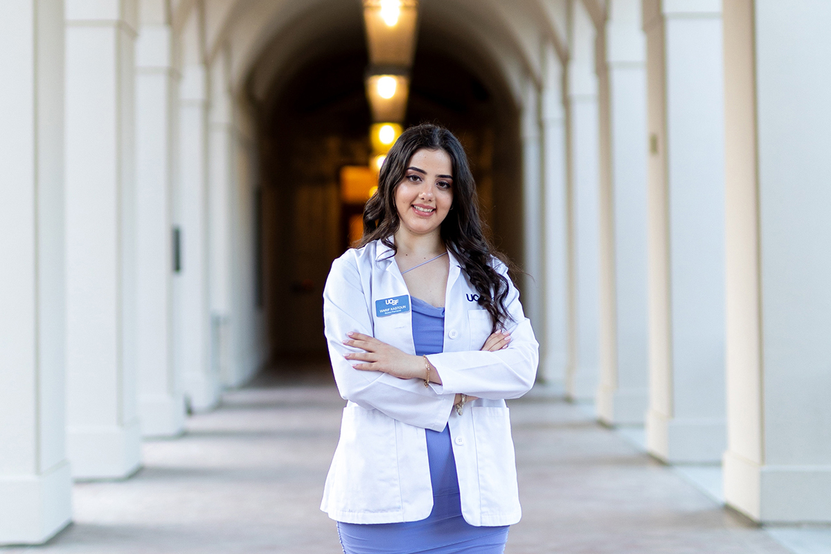 A young woman with long dark hair in a white coat poses surrounded by columns, arms crossed