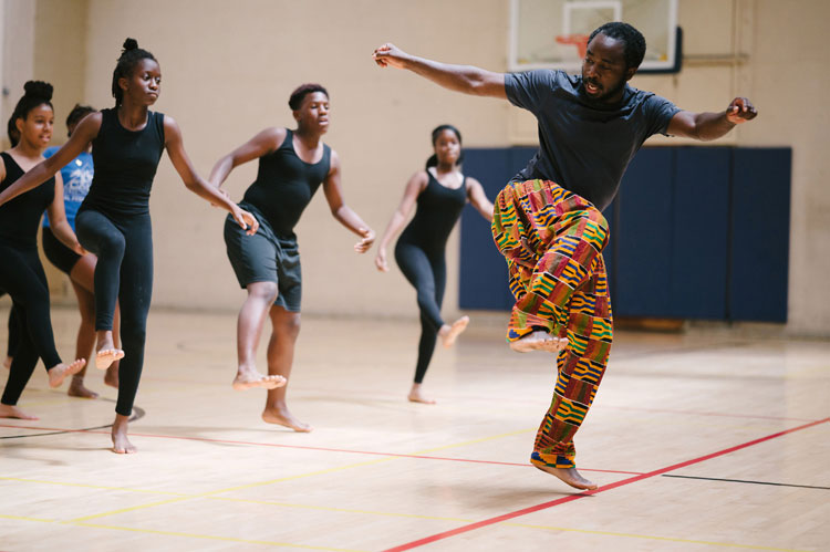A dance teacher in colorful pants demonstrates a kicking move while students follow along