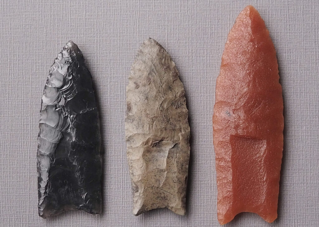 Three spear tips side by side on a linen-colored background. The left-most is black, the middle one ochre and the rightmost is salmon-colored.
