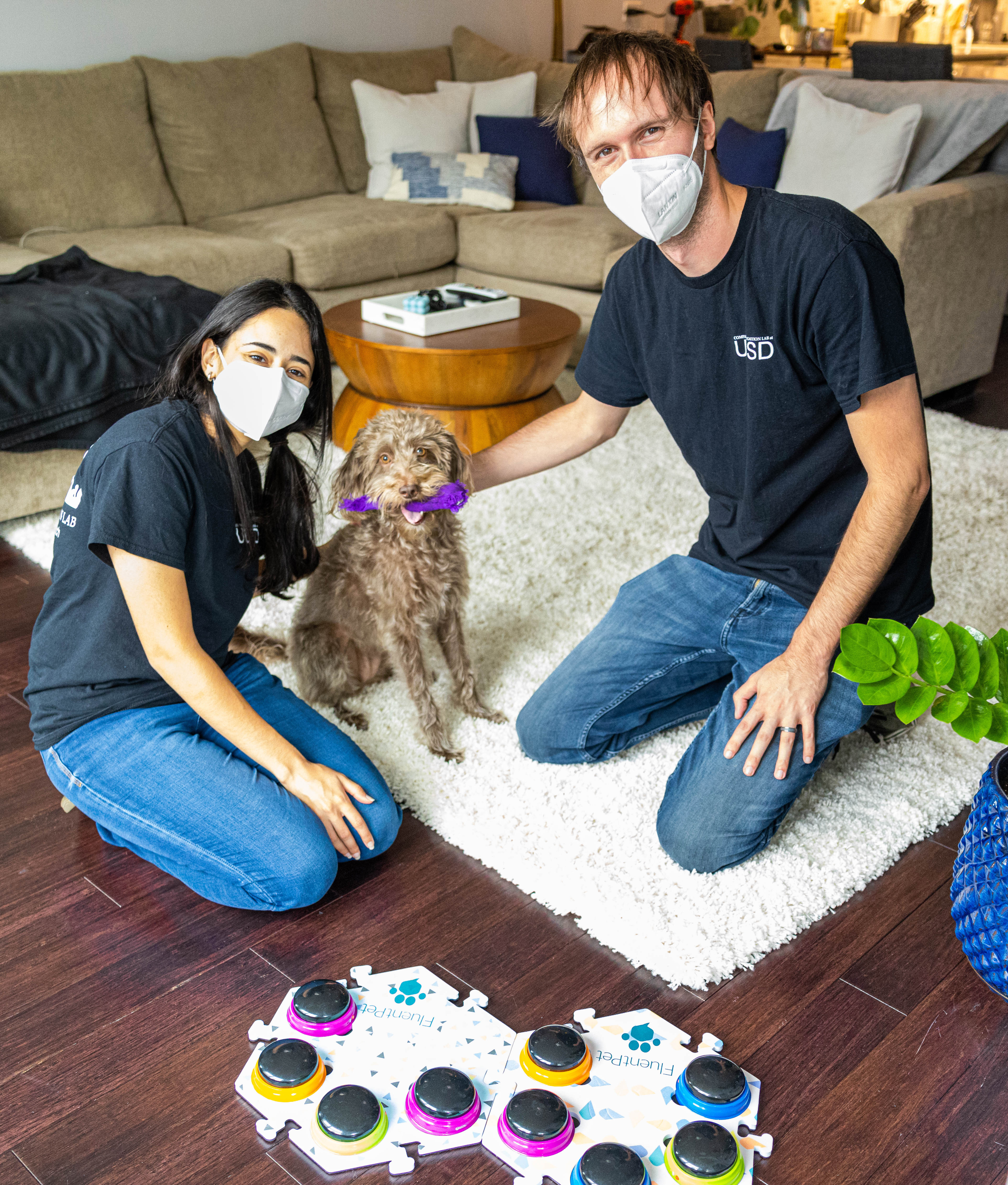 Two people wearing black T-shirts, jeans, and N95 face masks petting a small brown dog with a button soundboard in the foreground