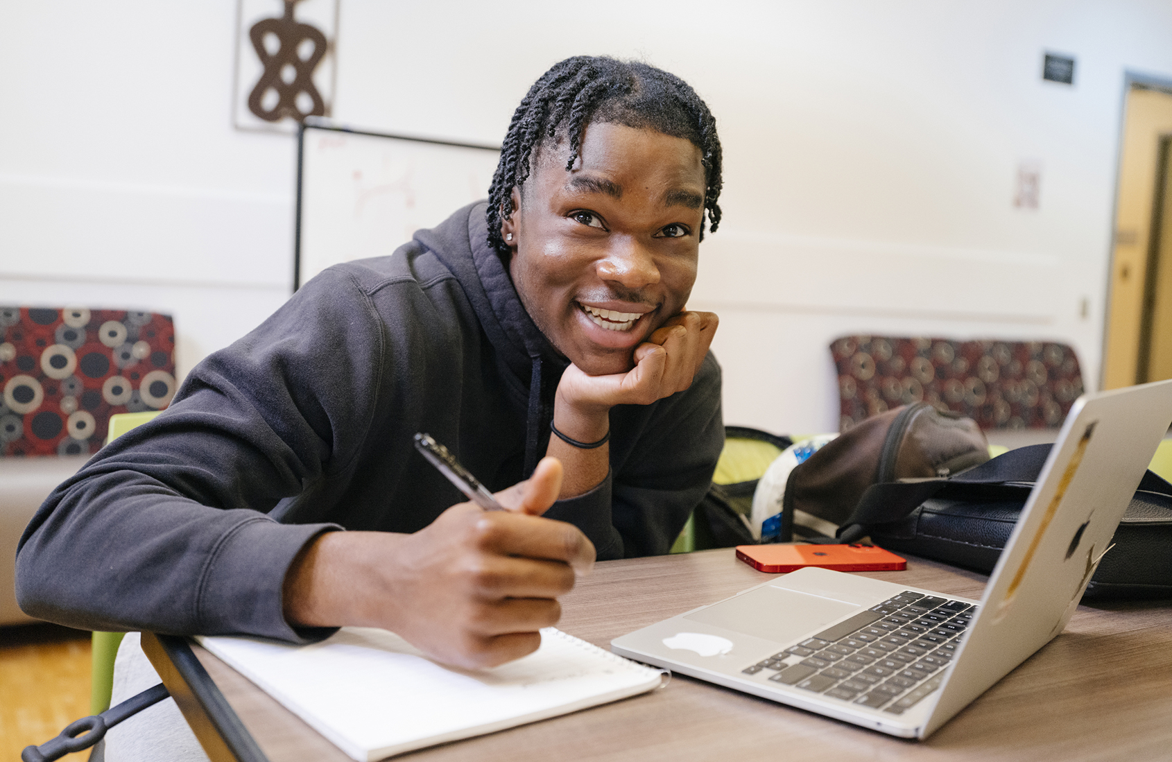 A college student sits in front of a laptop, chin propped on his hand, taking notes in a notebook with his other hand, looking up and the camera and smiling