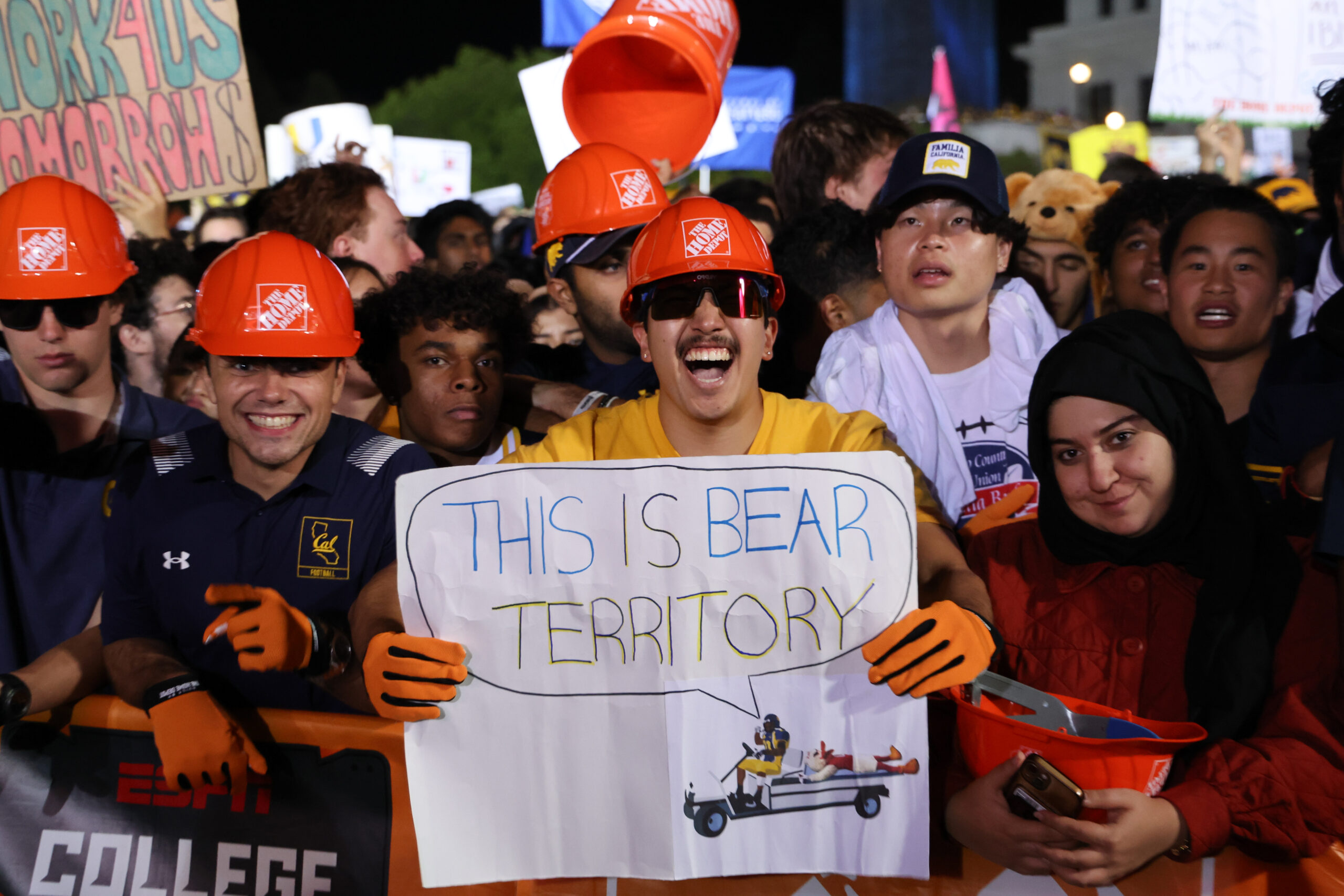 A group of students, some of whom are wearing orange Home Depot branded hard hats, scream at a camera, with one holdling a sign that says This is Bear Territory with a drawing of Marshawn Lynch in a golf cart, a woman in a headscarf next to him smiling 