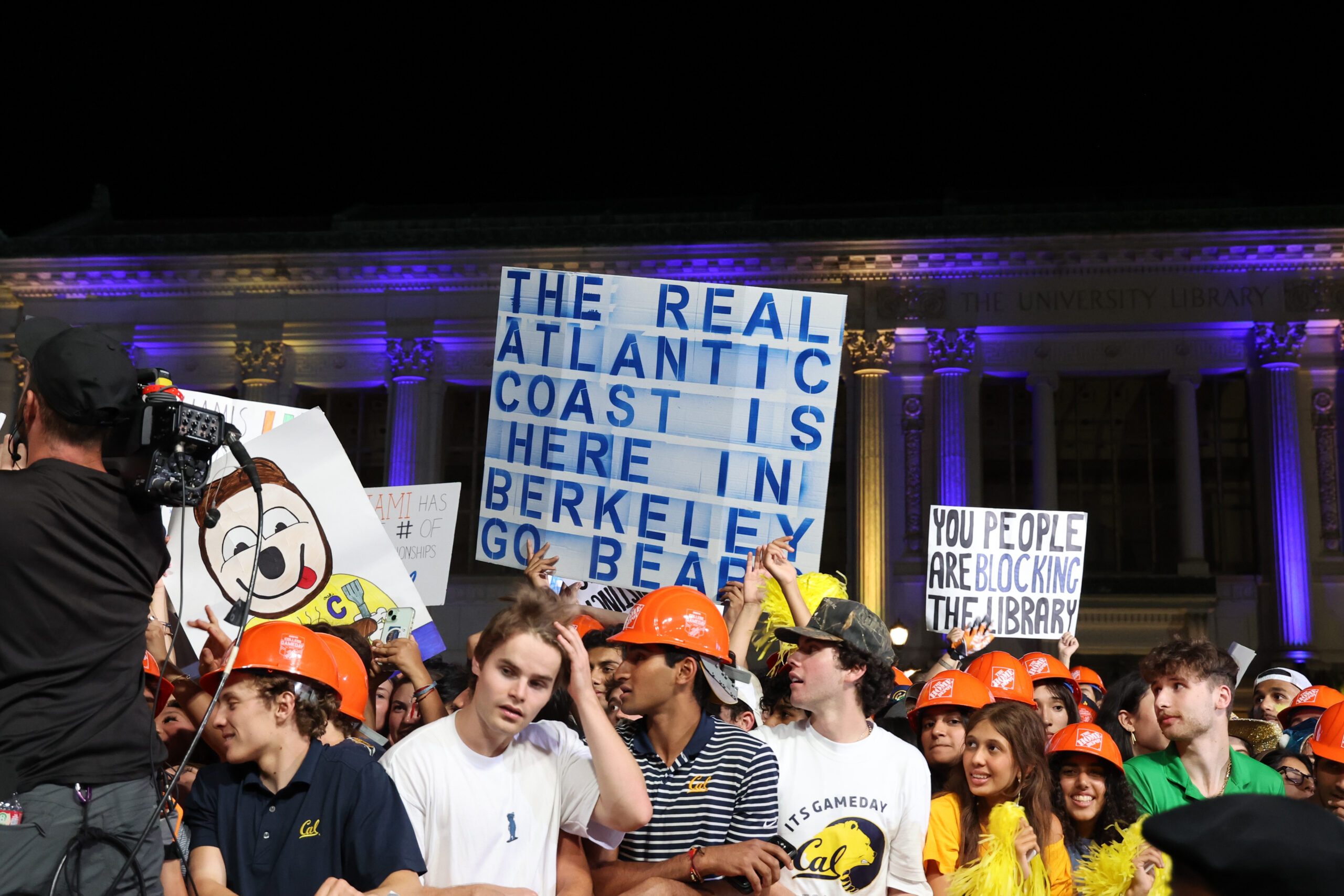 A group of students predawn, some of whom are wearing hard hats, some holding signs in front of Doe Library, for College GameDay