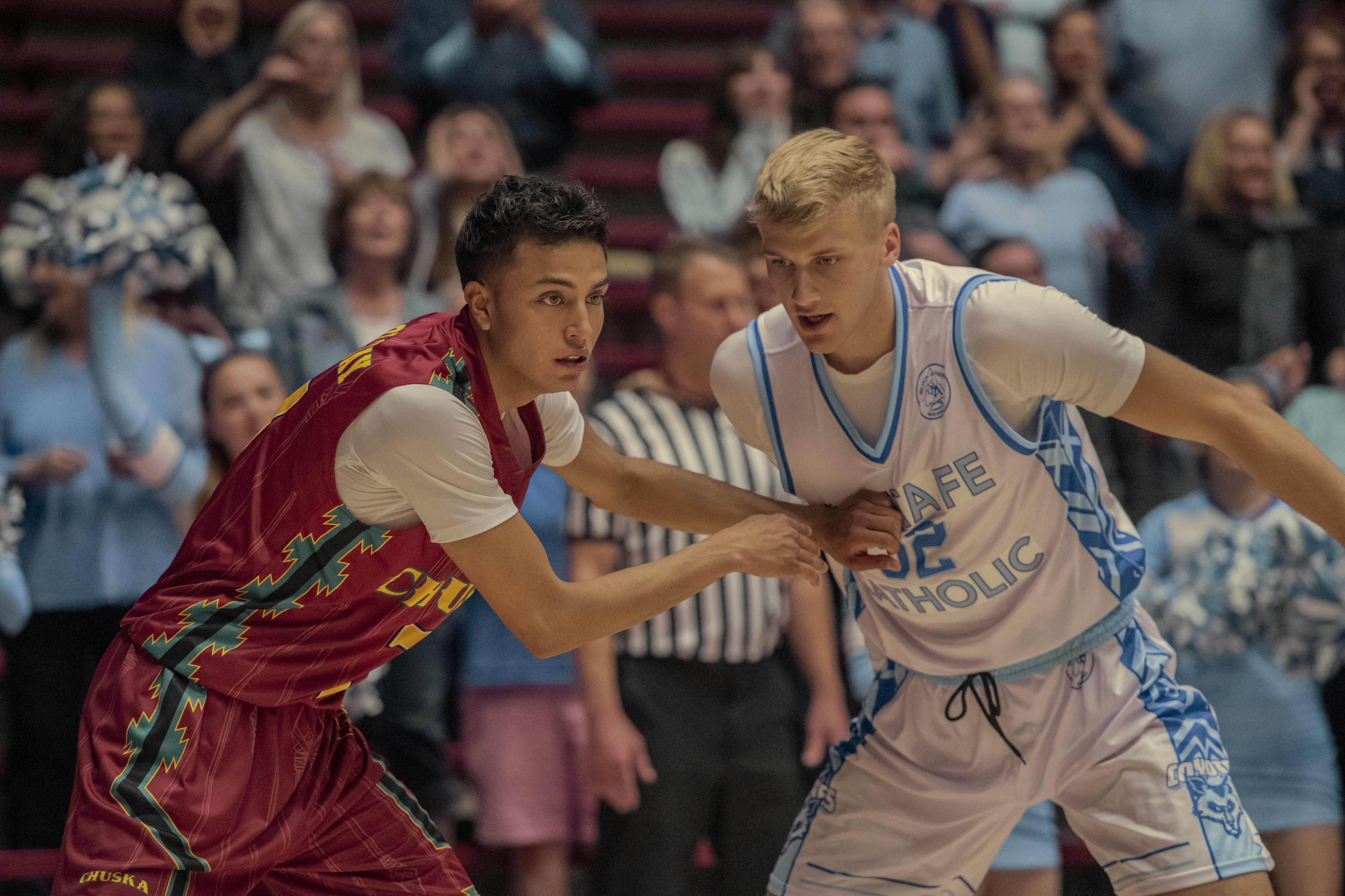 A young man with dark hair in a red basketball jersey and a young man with blonde hair in a white basketball jersey play in front of a crowd