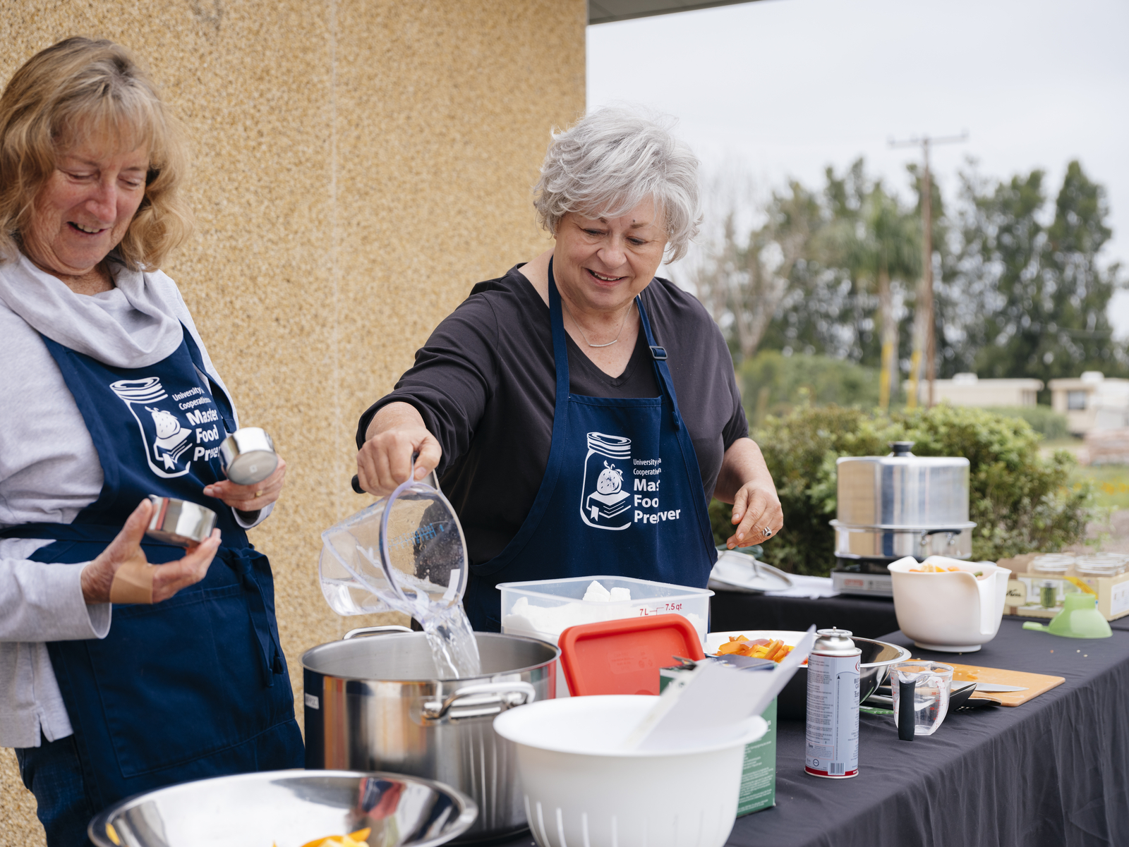 Two women in matching blue aprons with Master Food Preserver logos on them work at a table full of food preservation gear.