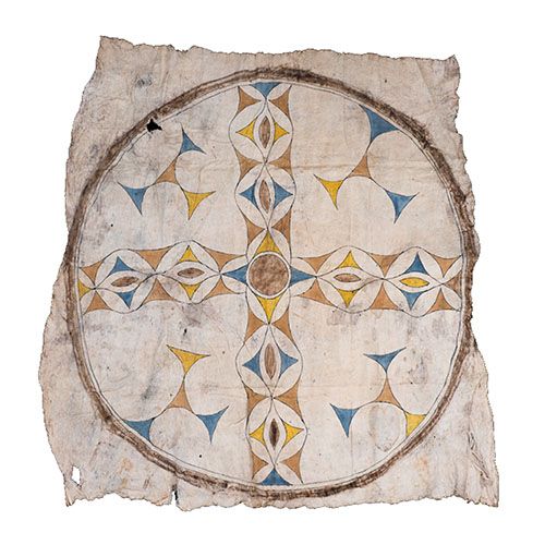 A Tikuna/Magüta item with a pattern of golds, browns, and blues in a cross-like shape, surrounded by a ring, with four three-pronged shapes in the space between right angles and the ring within