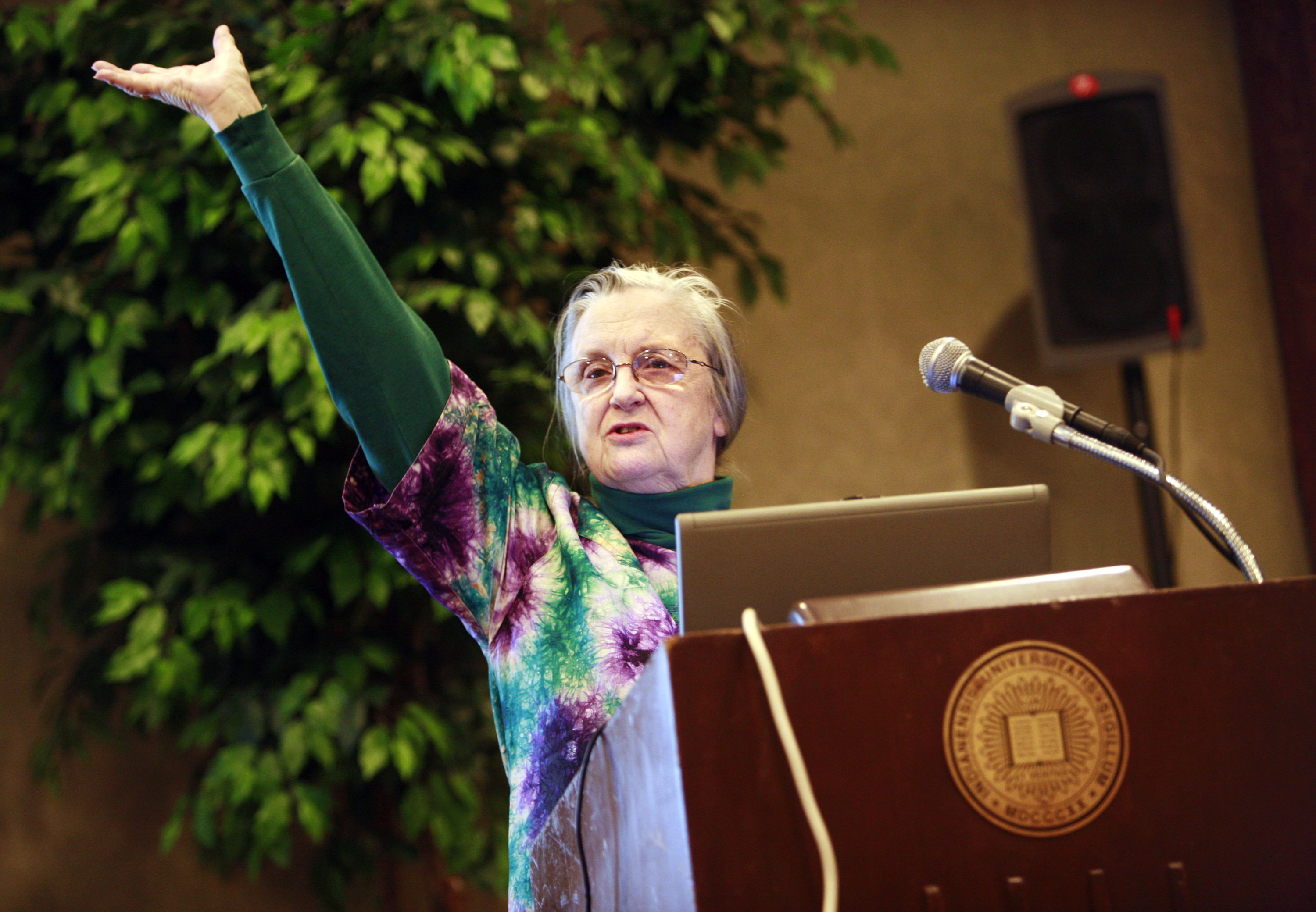 A woman stands before a podium, gesturing grandly with her arm in the air