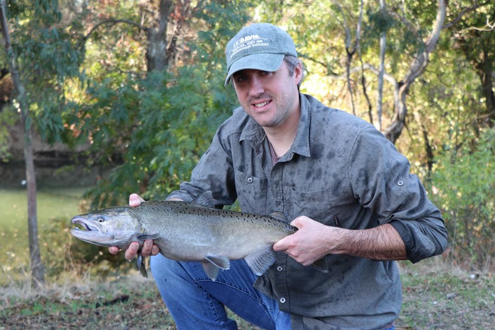 A man wearing a cap kneeling and holding a large fish, with trees and a creek in the background