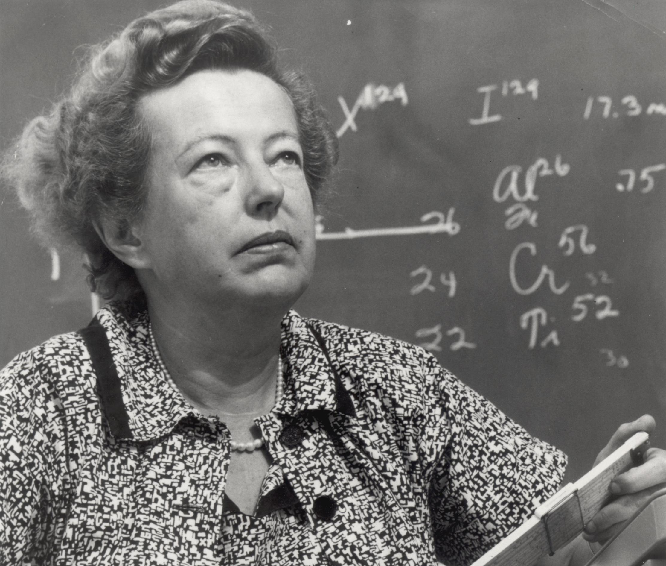 A vintage black and white photo of a woman in front of a blackboard marked in chalk with physics notations