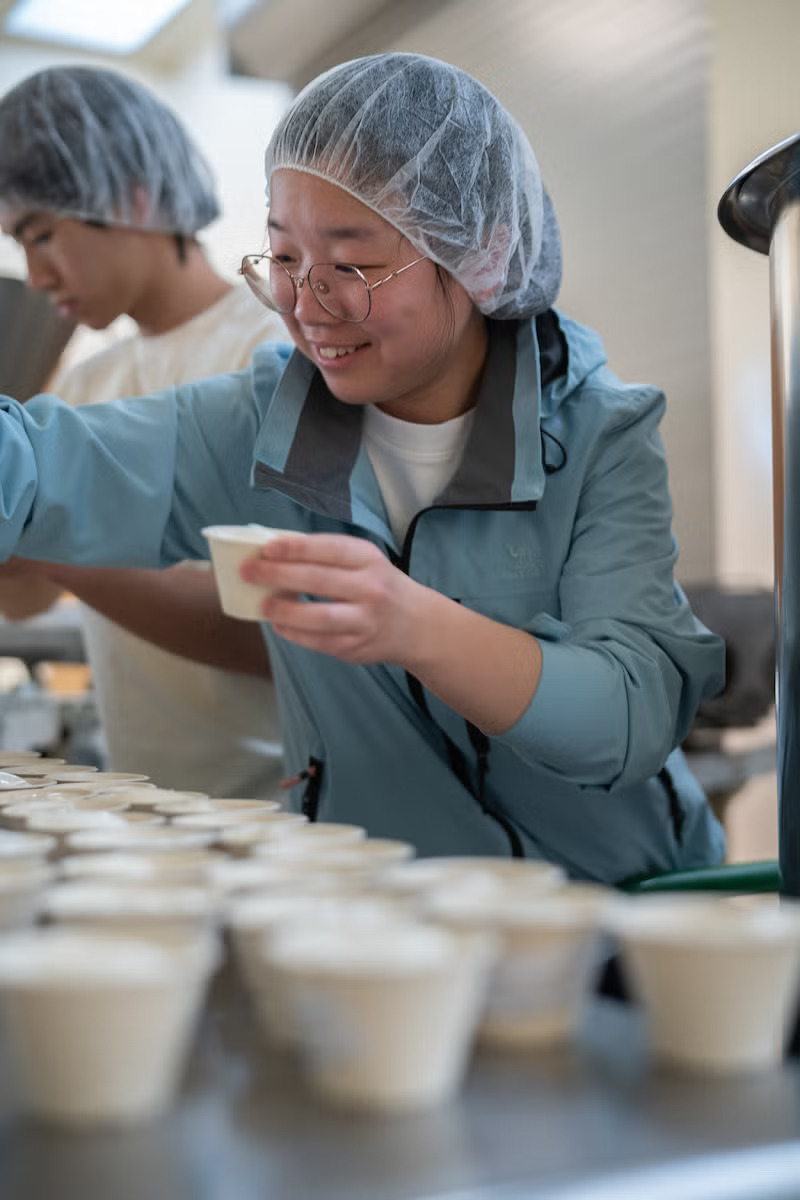 A young woman in glasses a hairnet and a light blue protective jacket smiles as she puts ice cream in cups