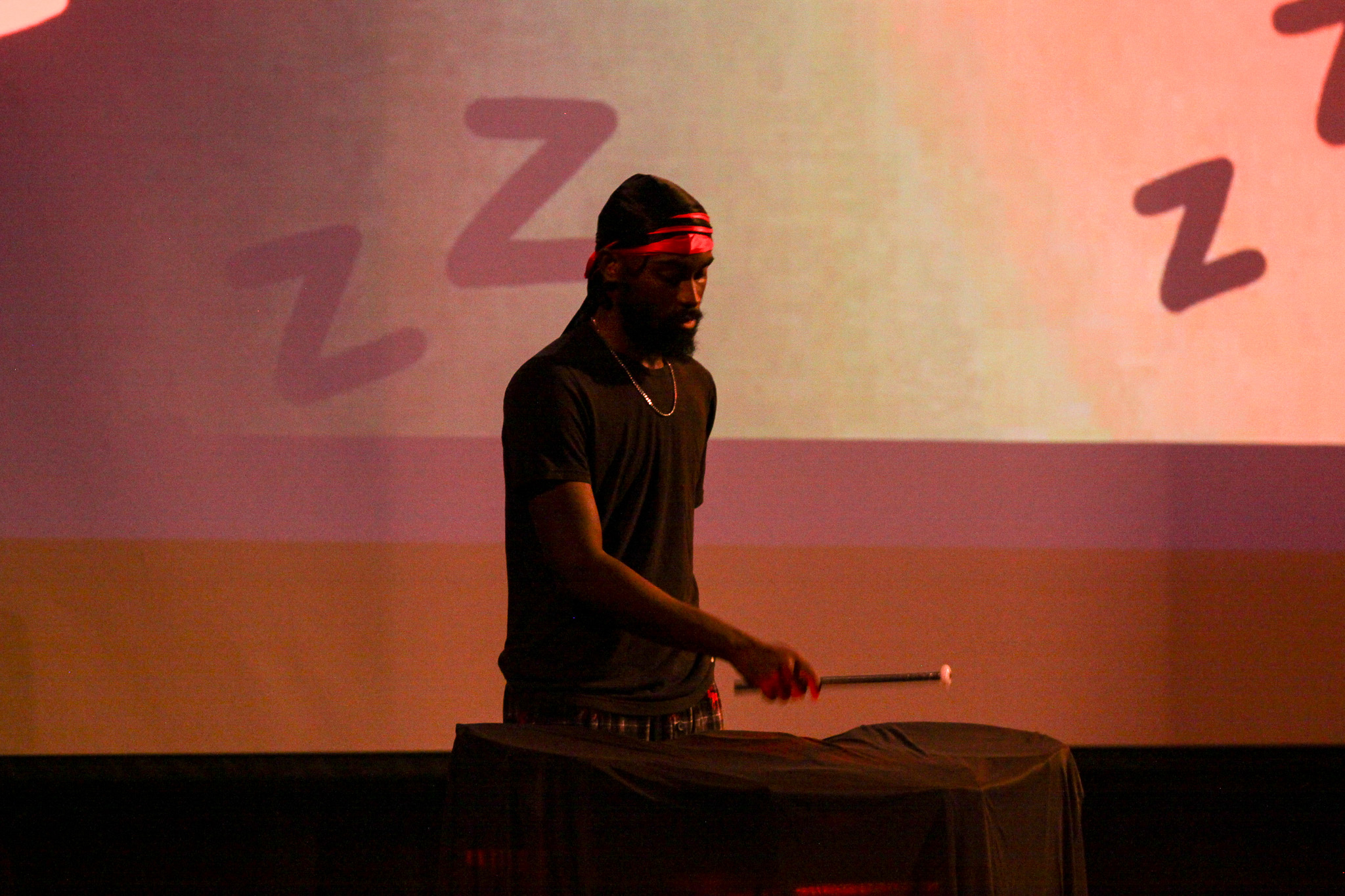 In a dramatically lit room, with Zs on the backdrop, a young man plays a drum