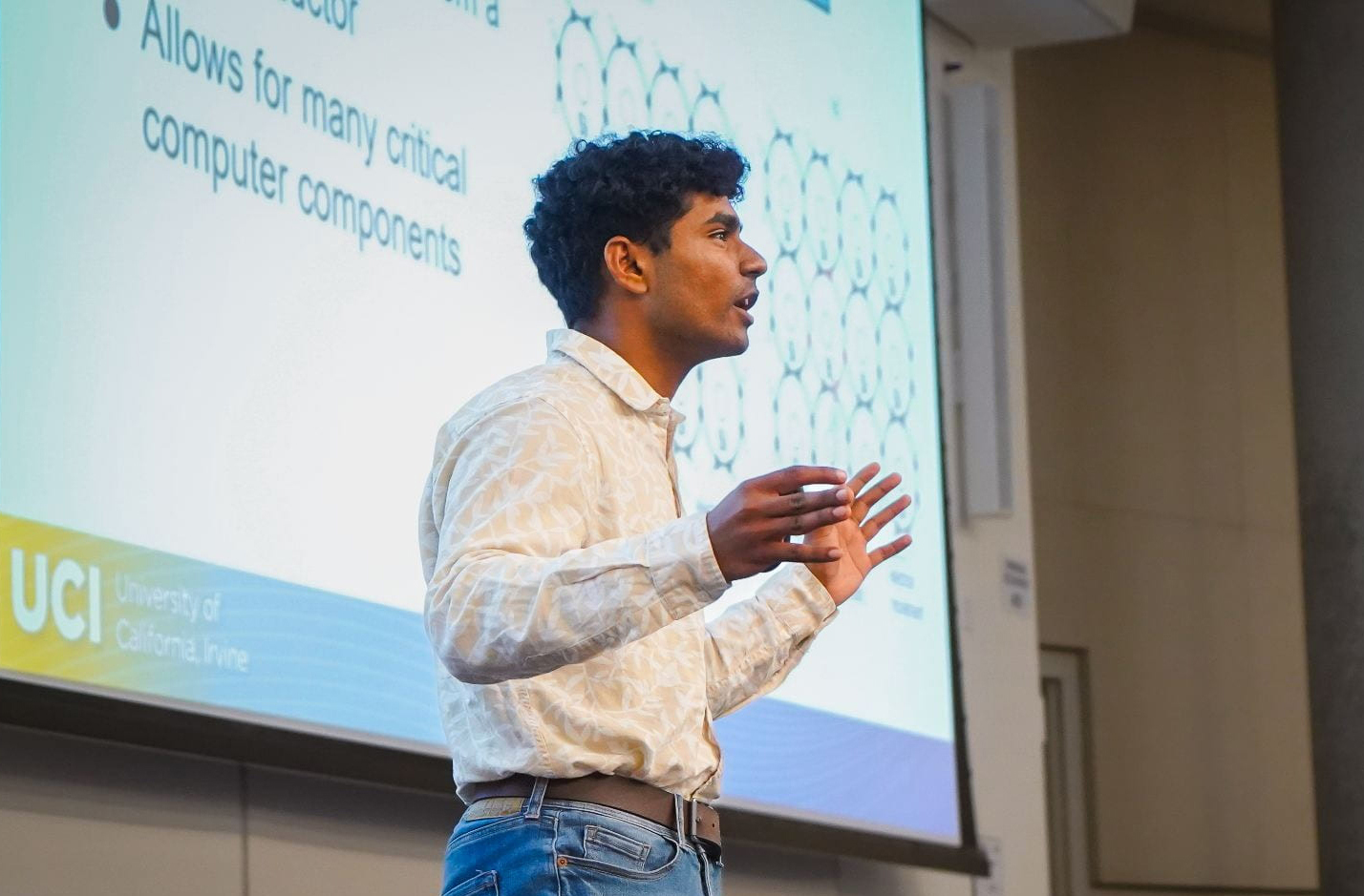 A young man in a white-collared shirt gives a talk in front of a large screen