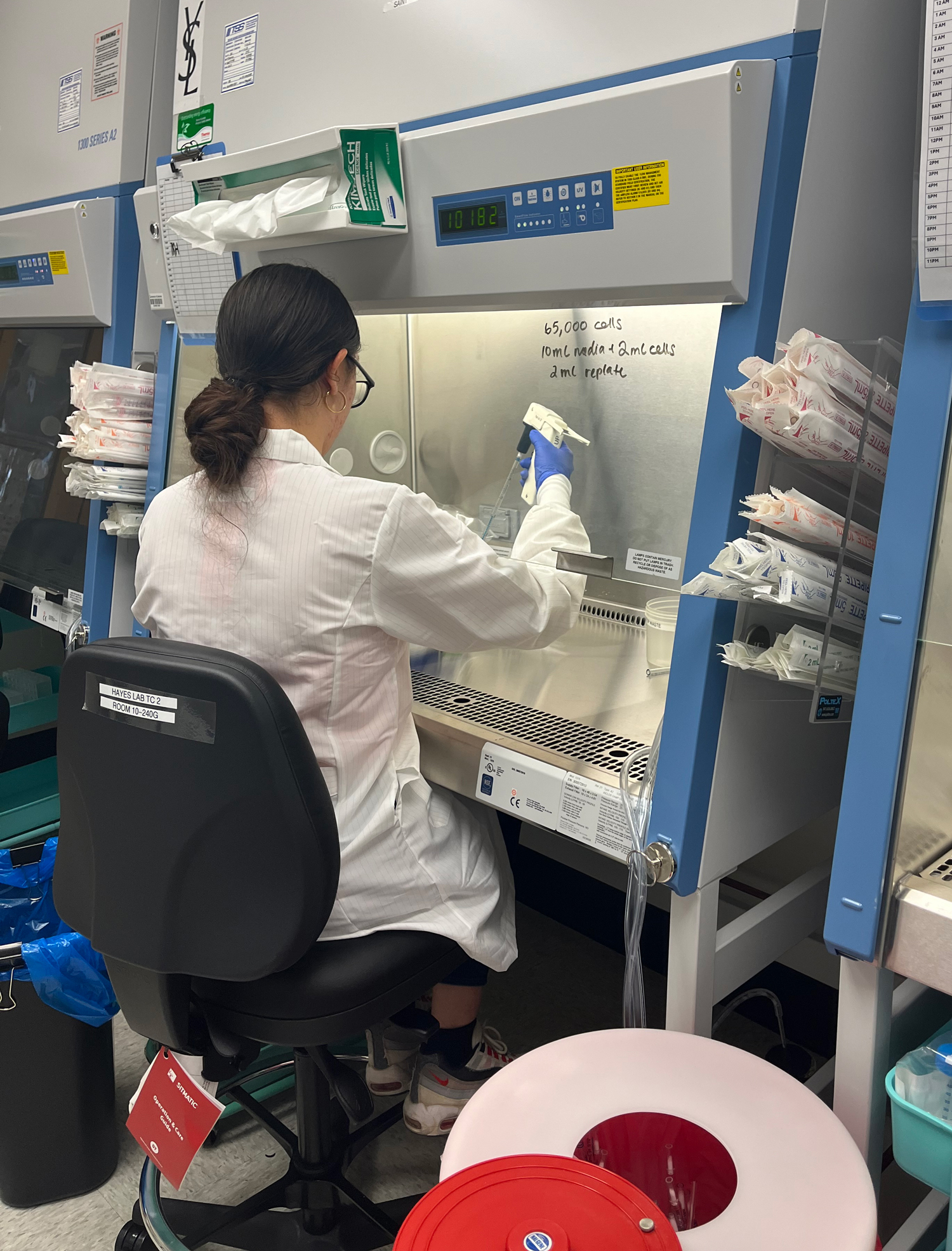 With her back to the camera, a young woman in a lab coat and gloves works at a technical-looking cube