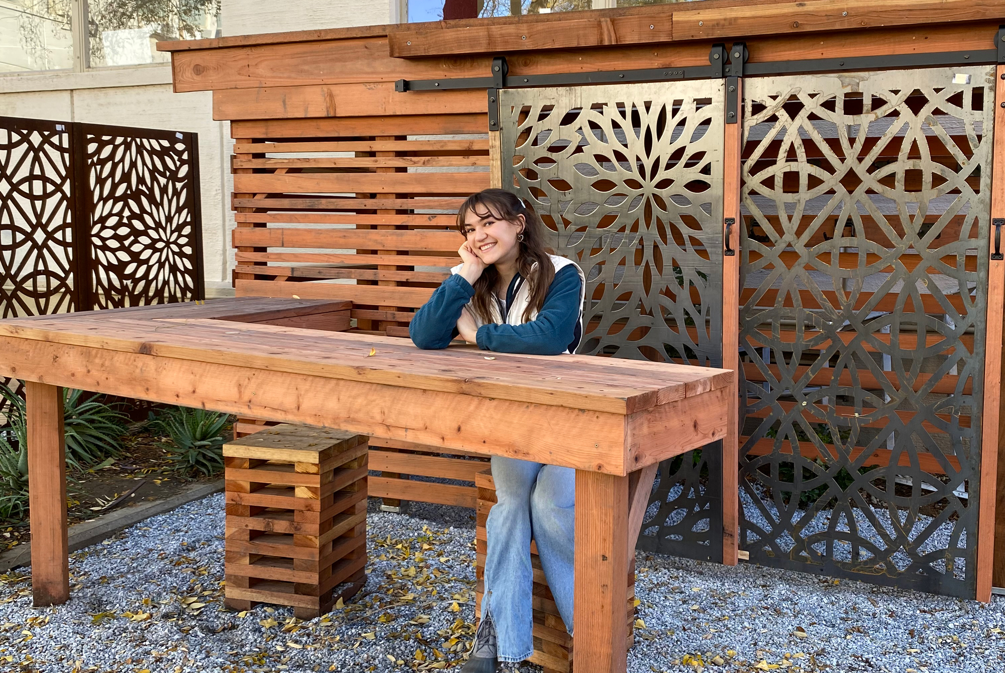 Young woman holds up her hand while leaning against a long wooden table in an ornately-designed semi-outdoor space featuring doors with wrought metal leaf designs