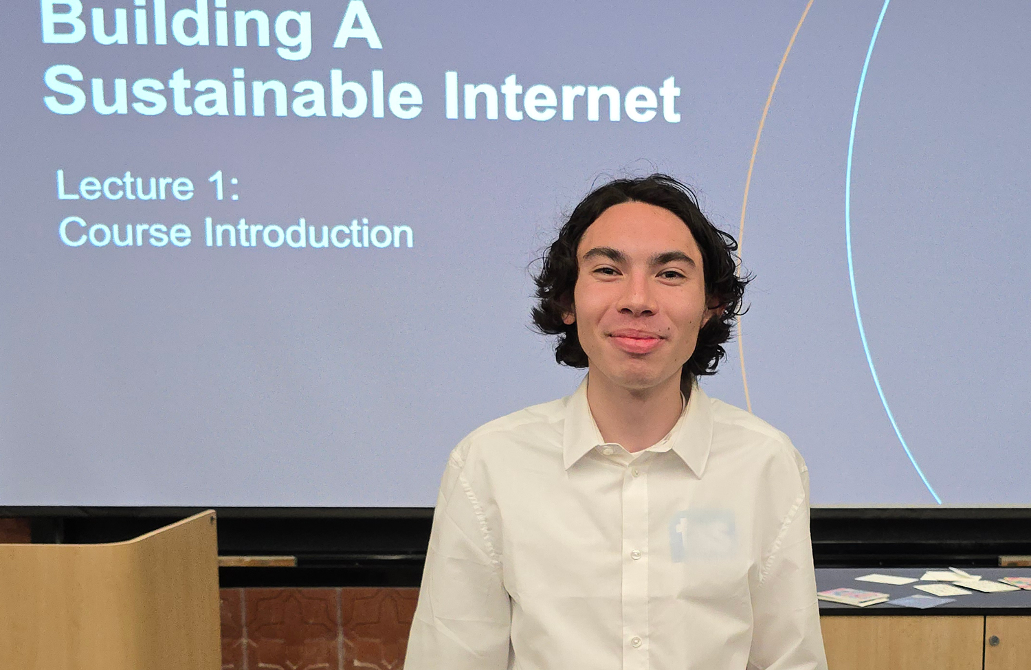 A young man smiles in a white collared shirt in front of a presentation slide that says Building a Sustainable Internet