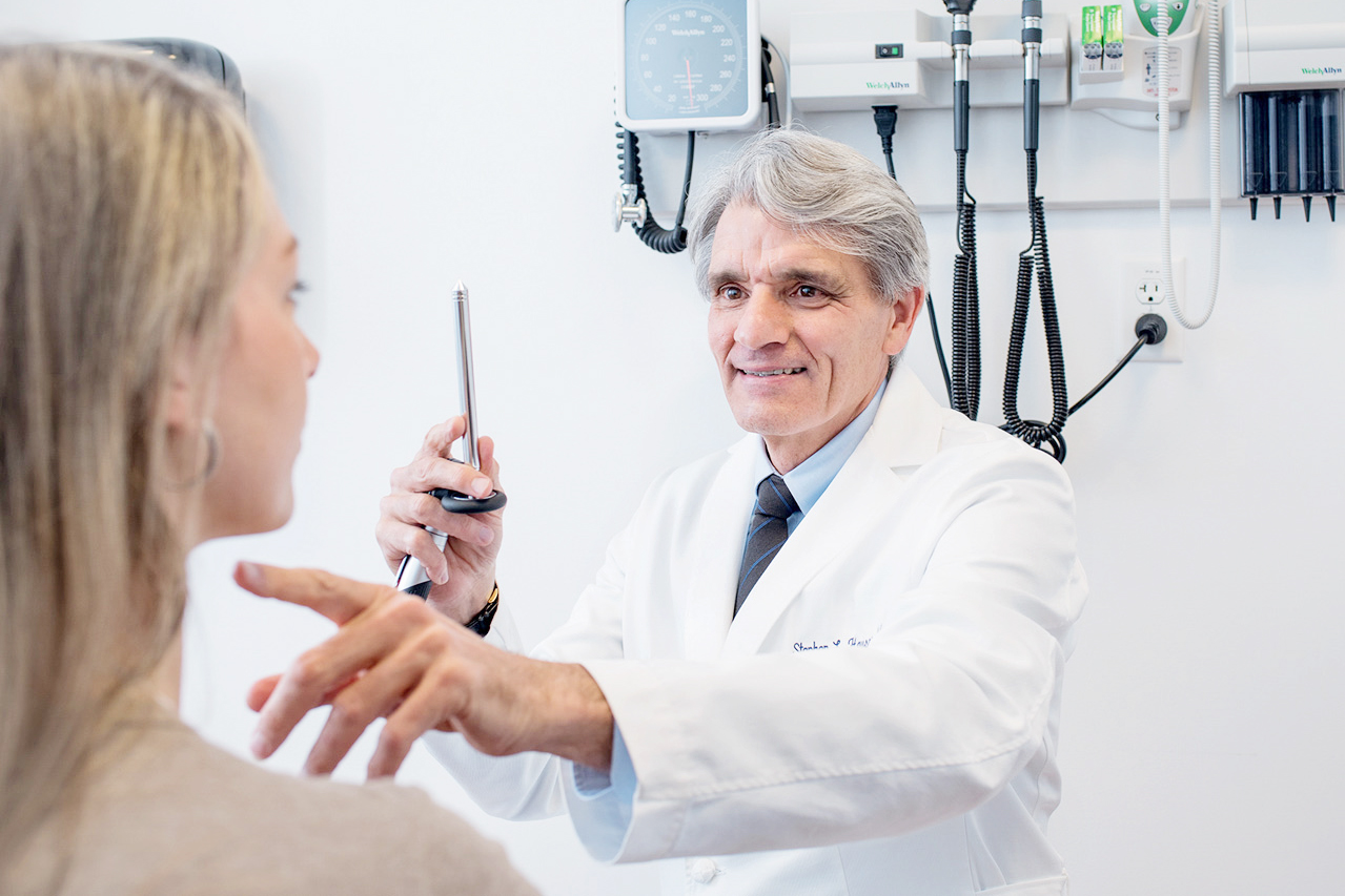 Doctor with white hair in a white doctor's coat points at a young patient's throat in a doctor's office