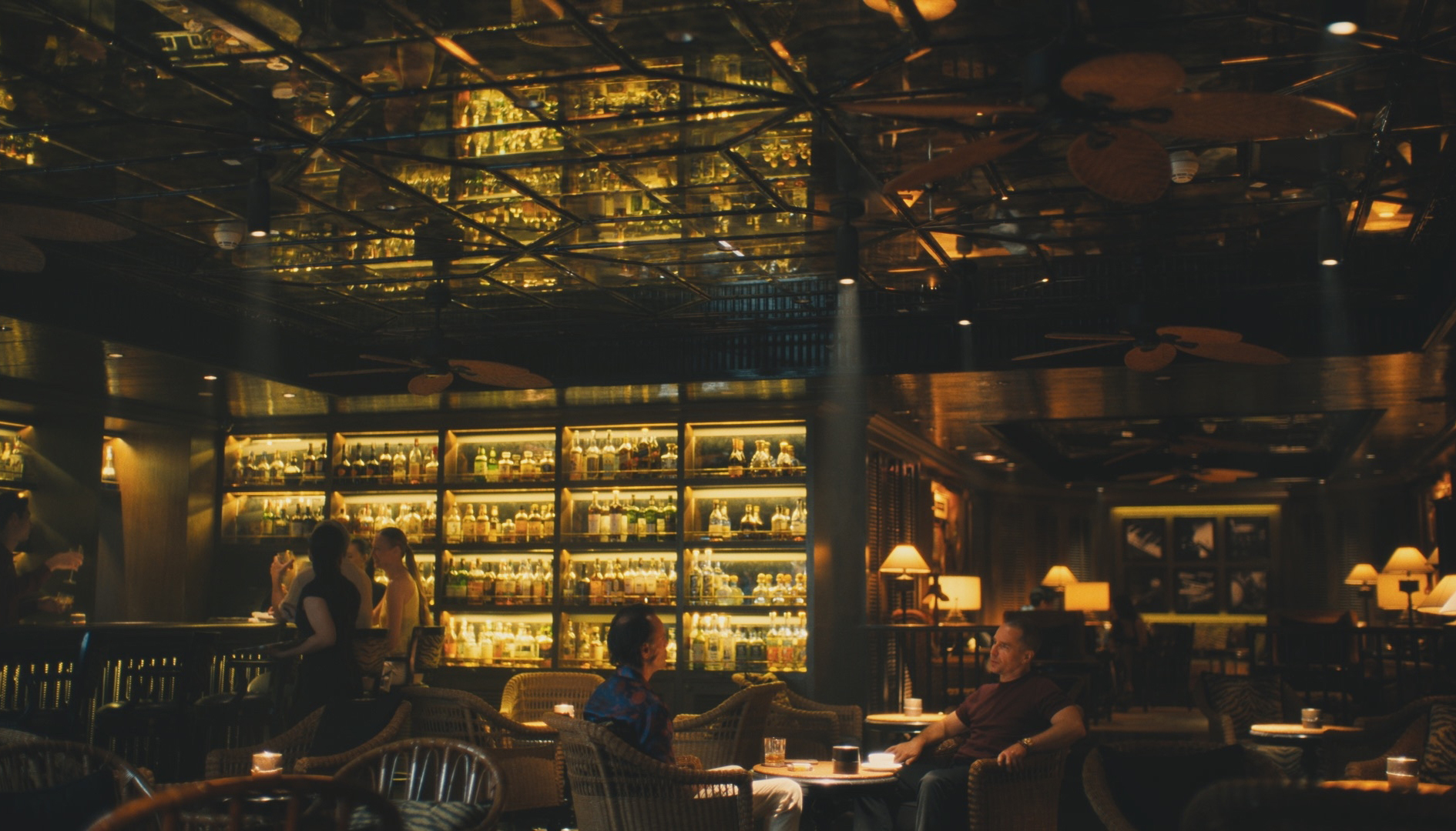 Two characters sit in a bar lounge with many mirrors and yellow light in a scene from The White Lotus