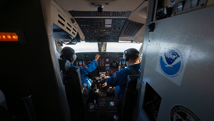 Two pilots in a cockpit of a jet, with the NOAA logo plastered on a bulkhead
