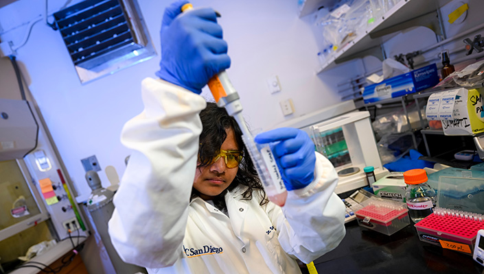 A researcher in a white lab coat squirts something from a bottle into a beaker in a lab.