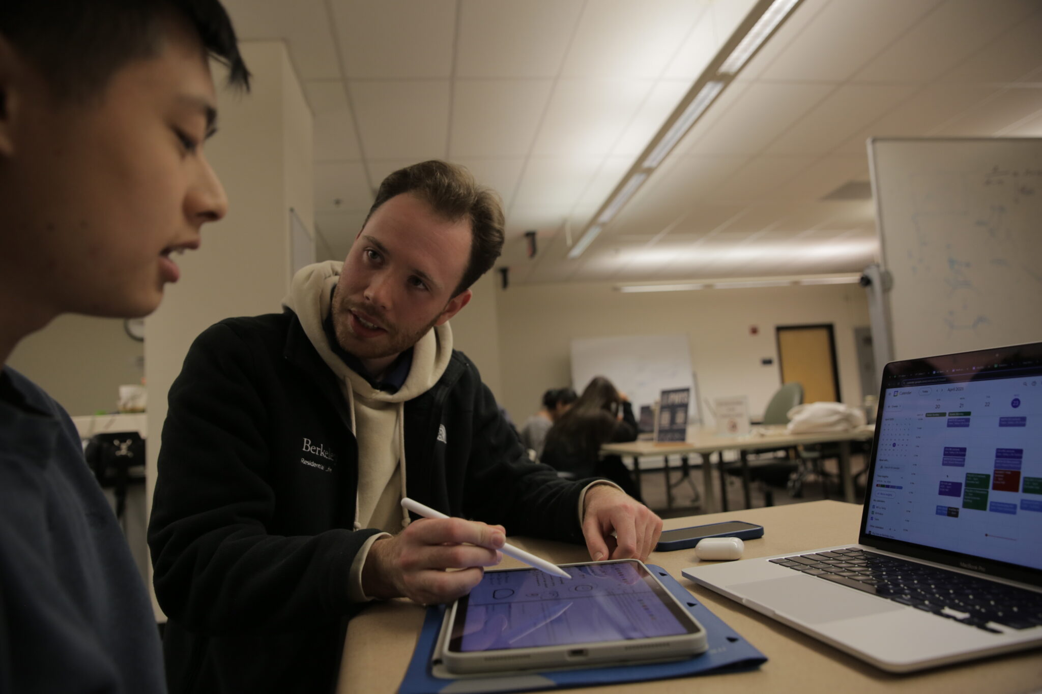 A young man holds stylus and looks at another student as they consider a laptop with a calendar and a tablet that the other student is looking at