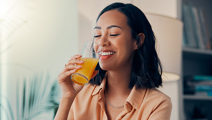 A woman smiles down at a glass full of orange juice as she lifts it to her mouth