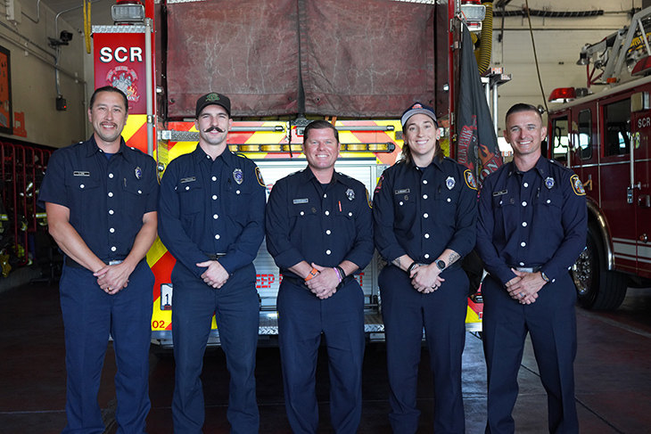 Five firefighters in blue uniforms pose in a firehouse in front of a truck