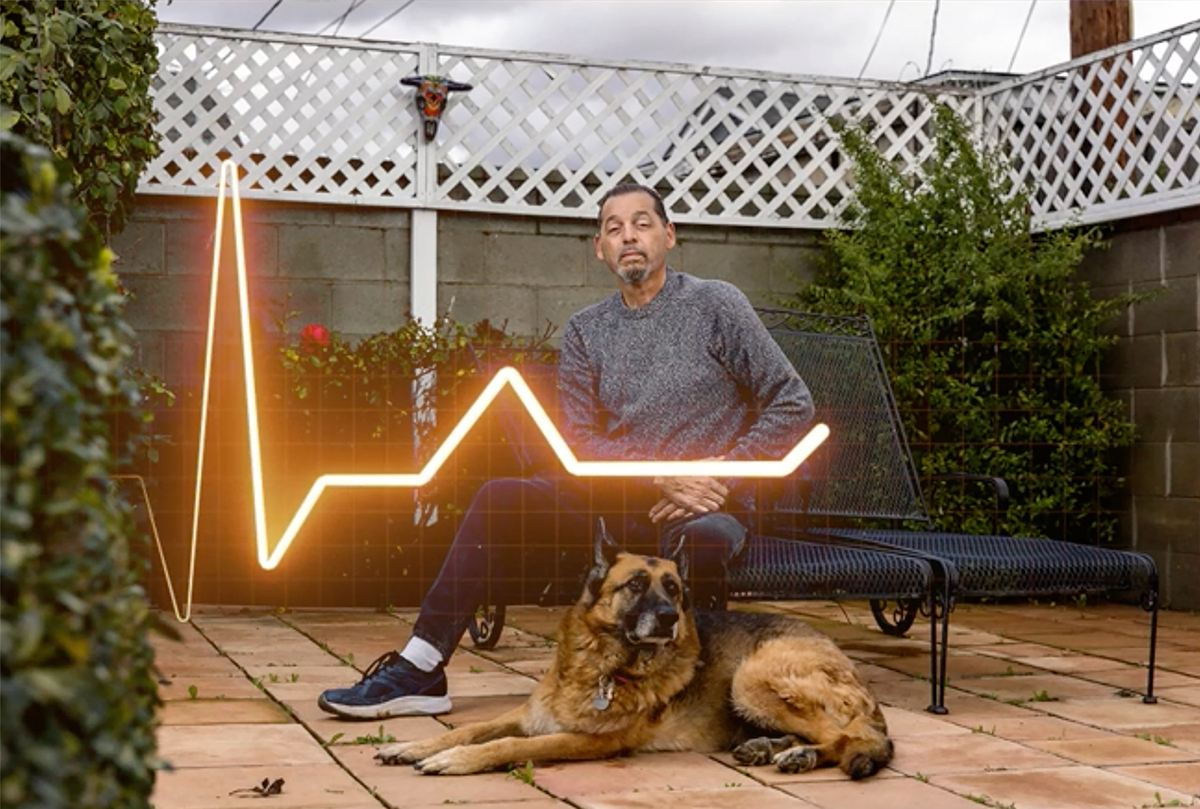 A man sits on a patio bench next to a German shepherd with an EKG line overlaid on the photo