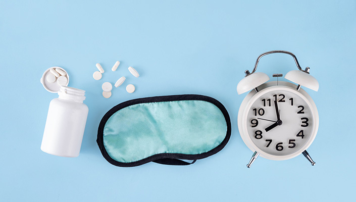 A bottle of pills next to a sleep mask next to an alarm clock on a light blue background