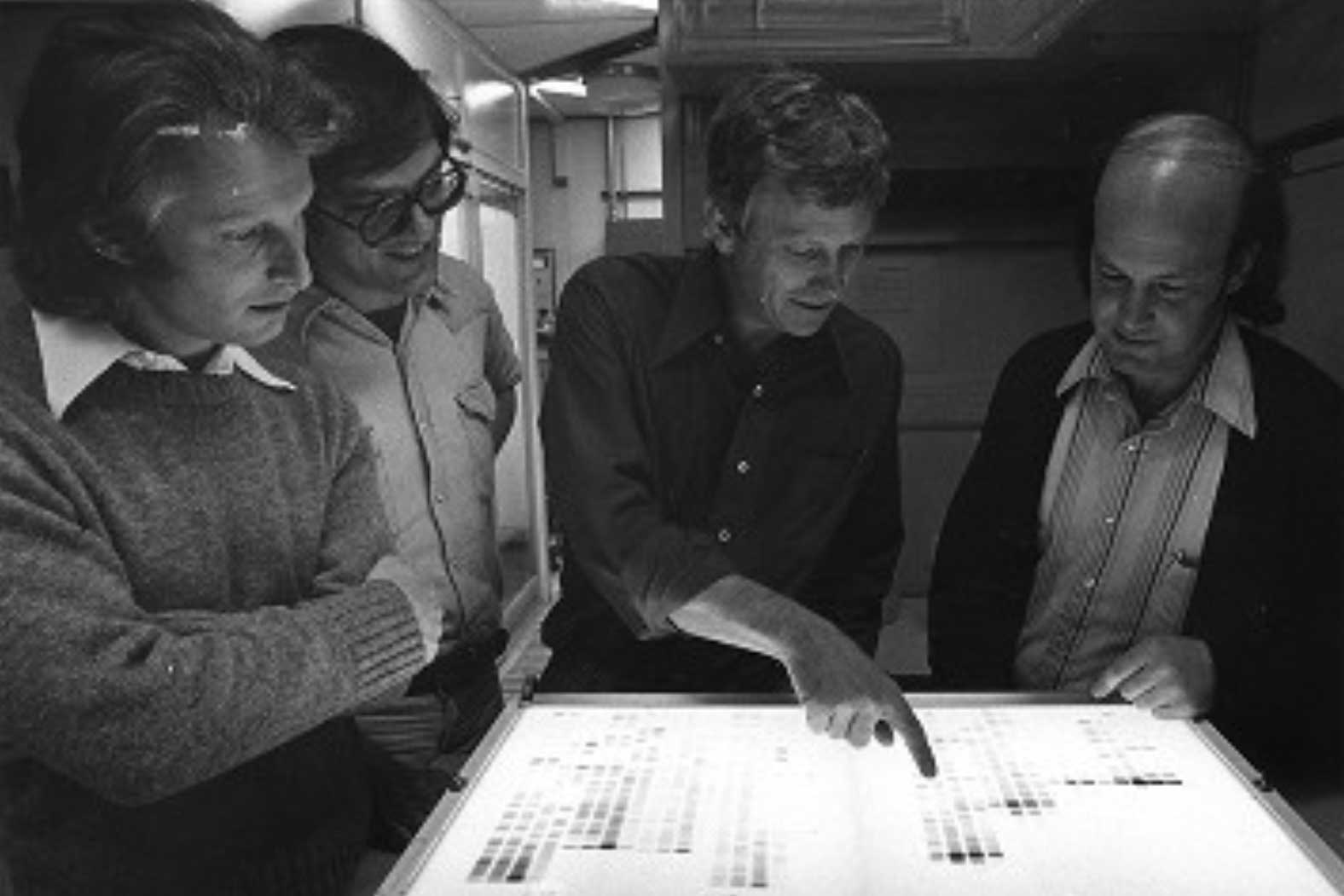 In a black and white photo four men look, one pointing, at a medical image