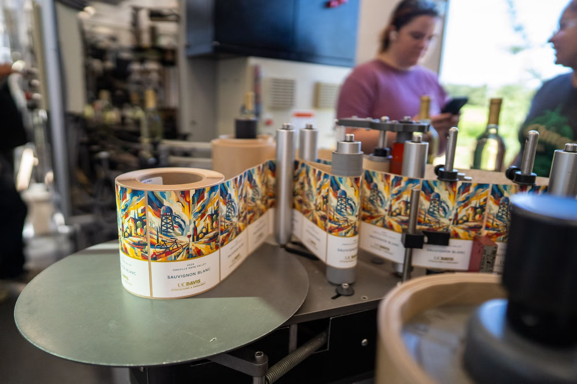 Colorful wine label images of UC Davis campus being affixed on wine bottles, with two people in the background