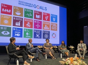 In front of a large projected image of colorful blocks, a group of five people sit in a panel, talking