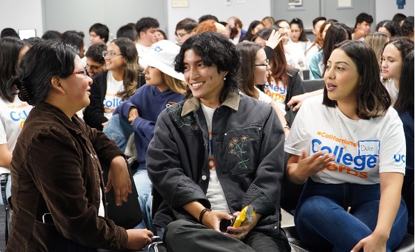 A young man, center, talks to someone to his right, while someone in a College Corps T-shirt is also talking to the same person, in a photo with a crowd behind