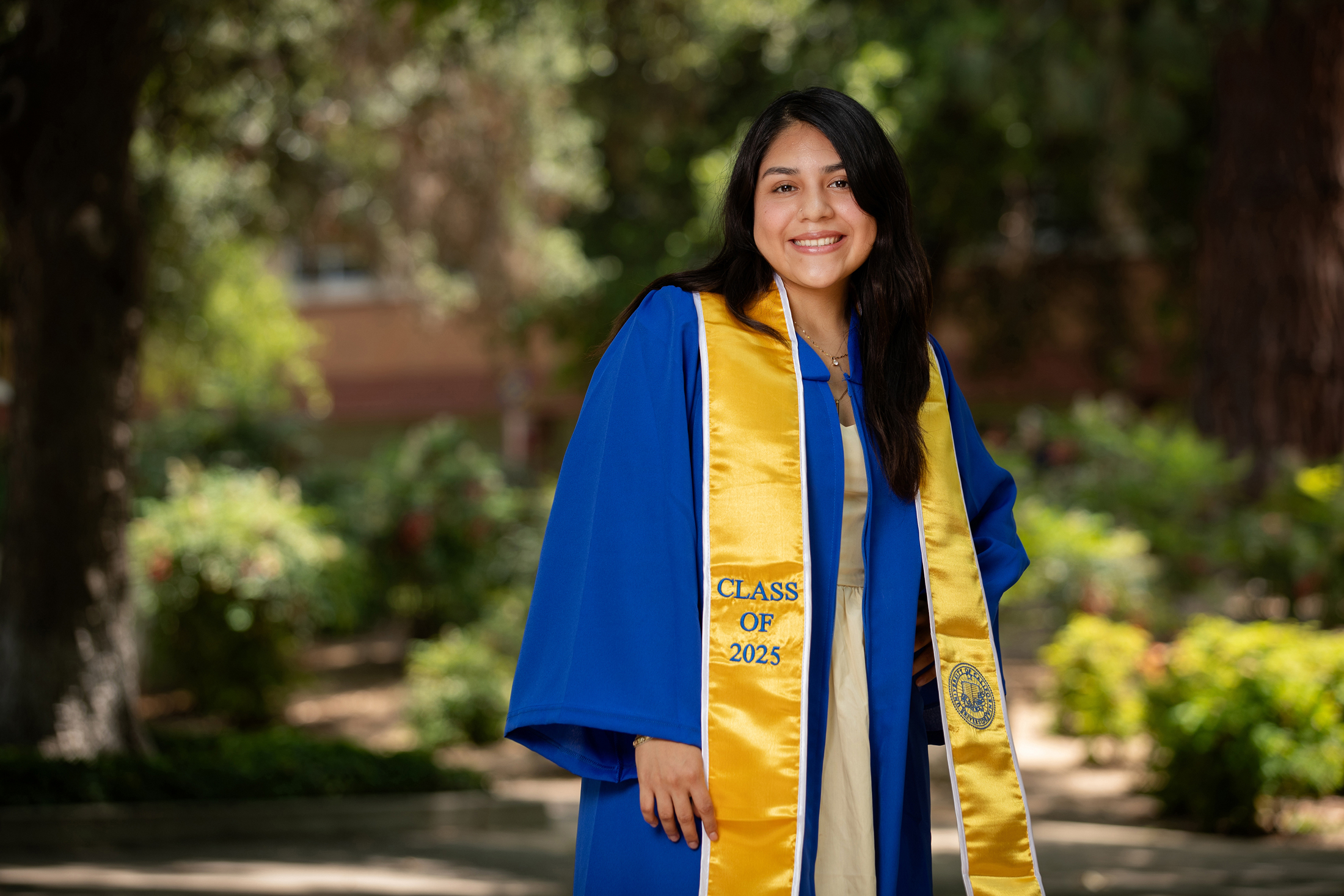 Young woman graduate with long black hair and a blue gown with yellow stole smiles on the UC Riverside campus