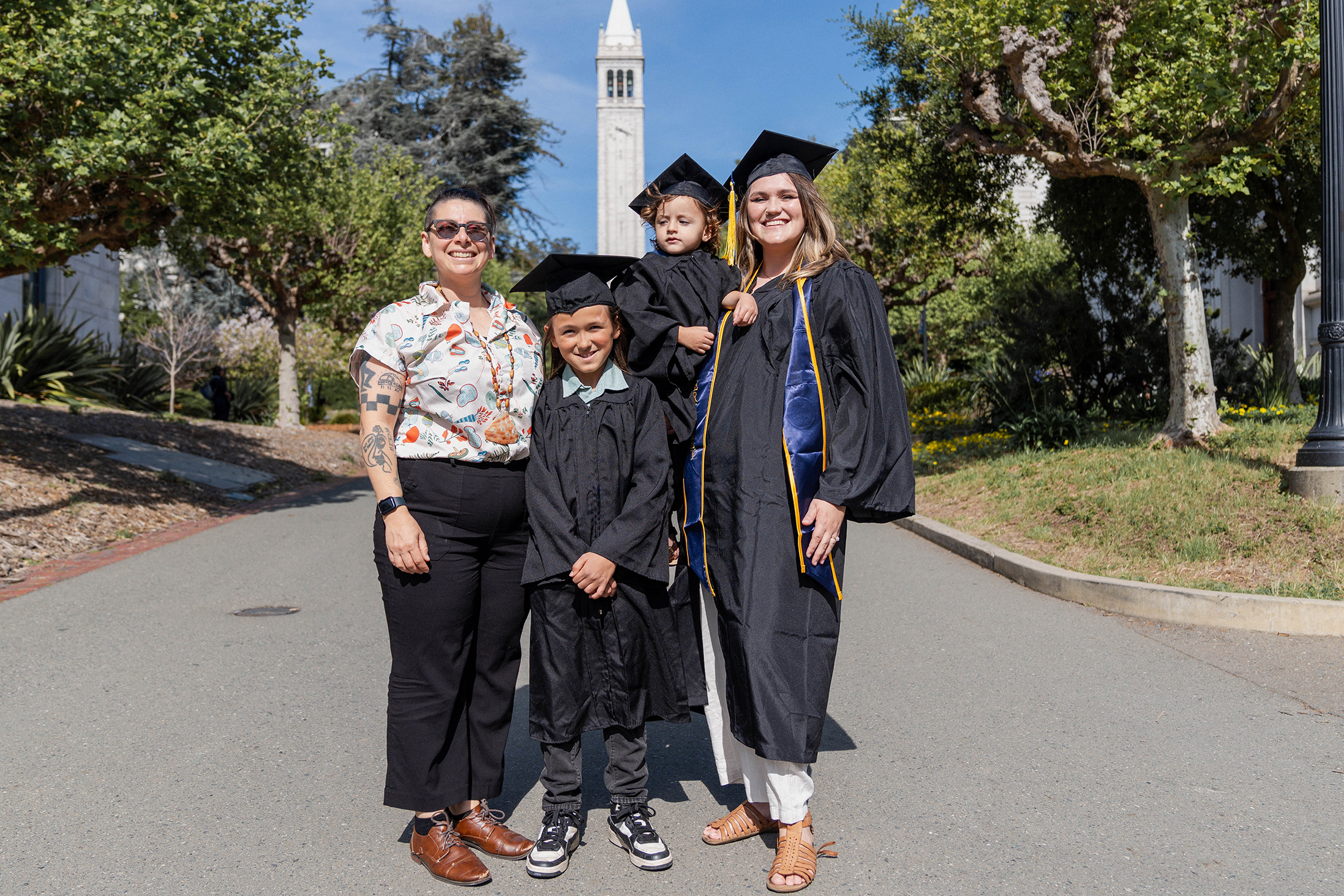 A family of four, two children, stand in front of the Campanile on the Berkeley campus, the two children and the woman on the right in cap and gown