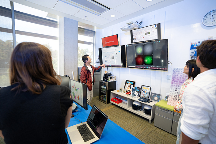 A few people stand in a room with many large monitors; someone close to one monitor points at it