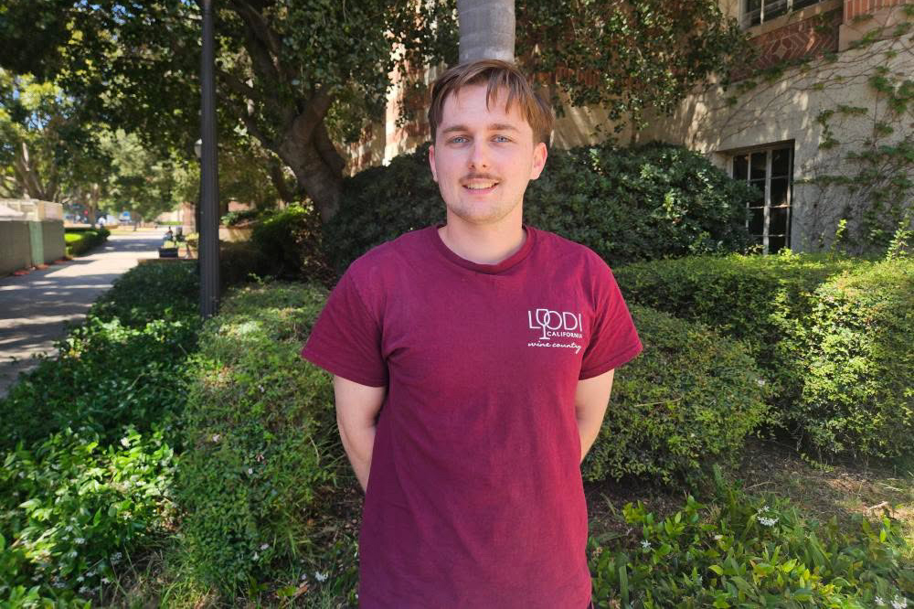 Young man with mustache smiles at camera in front of leafy area on UCLA campus