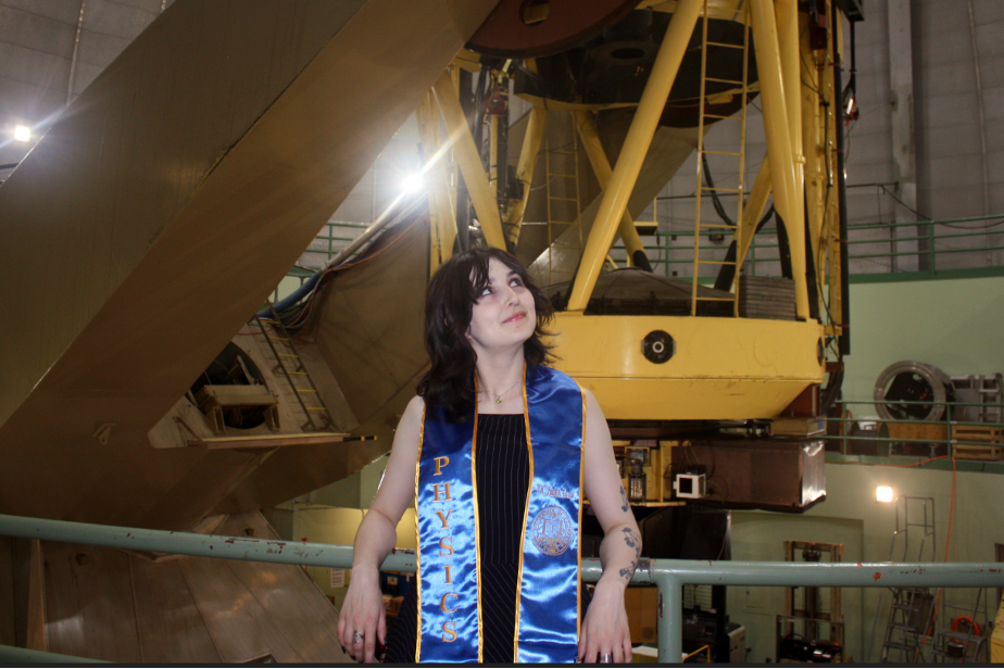 Young woman graduate with long black hair and a stole poses in front of, and while looking up, at a large piece of equipment