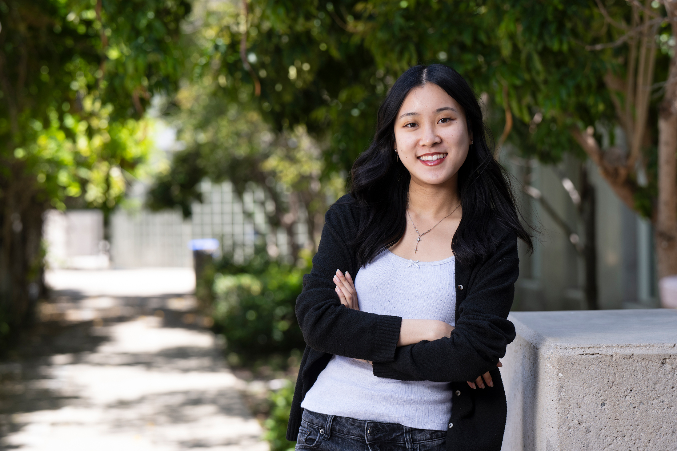 A woman in a cardigan crosses her arms and smiles, standing outside on a leafy sidewalk path at UC Irvine