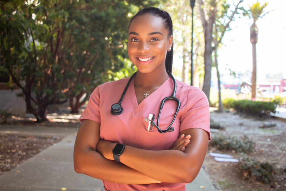 A woman in a pink nursing scrub with a stethoscope around her neck smiles at the camera, arms crossed
