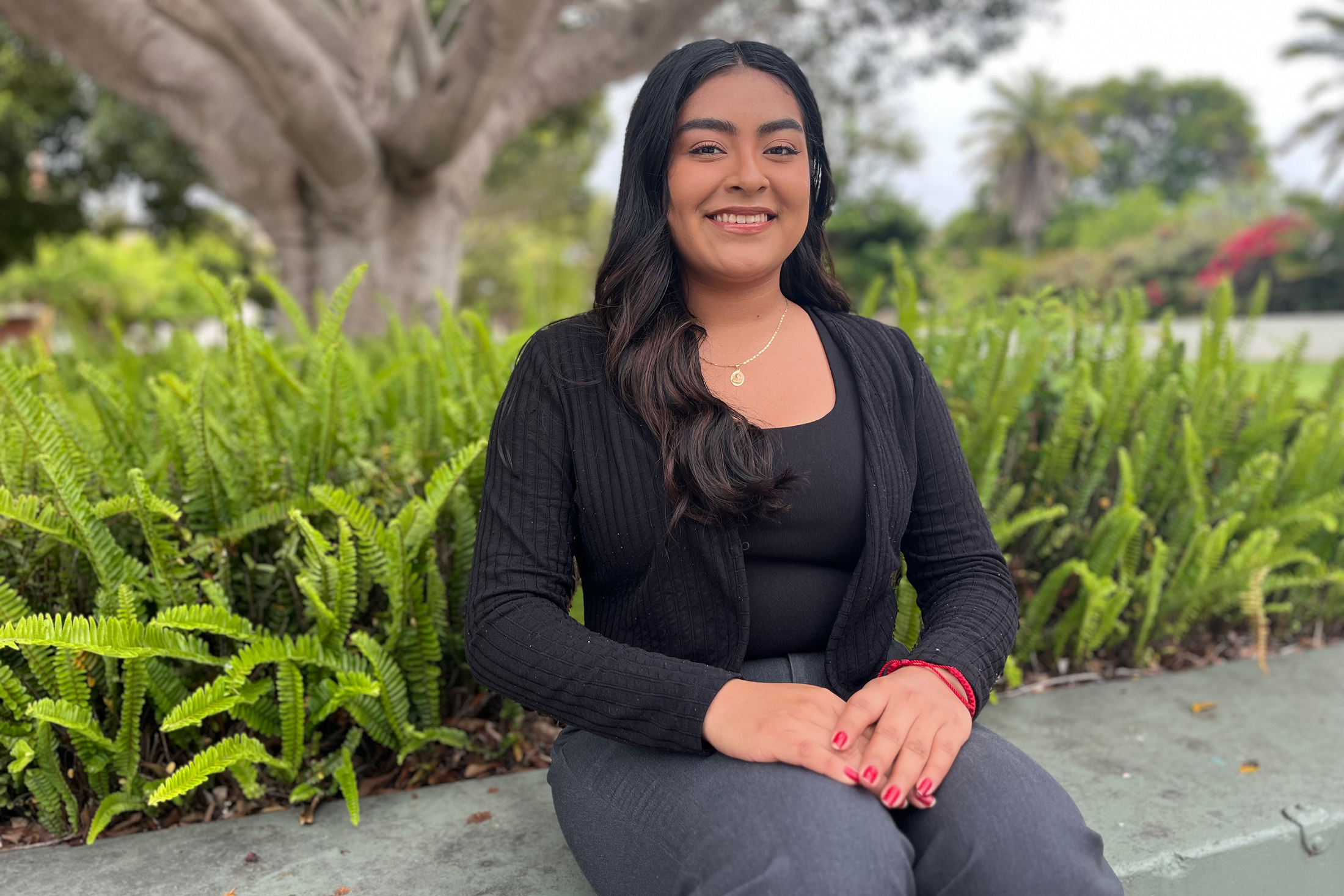 A woman in a black cardigan and black shirt smiles, seated outside at UC Santa Barbara in front of some greenery