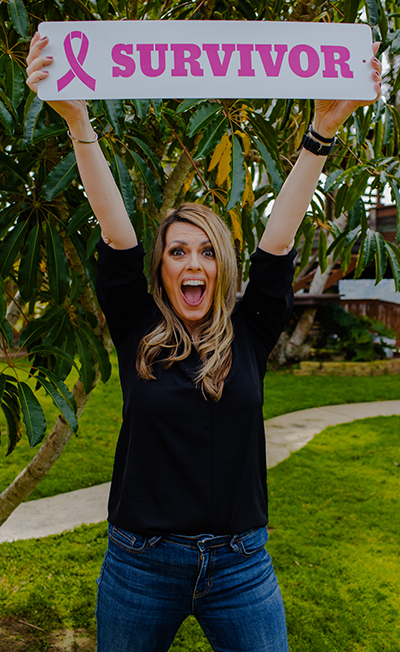 A woman with long curly blonde hair holds up a banner that says SURVIVOR with a pink ribbon next to it in excitement